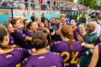A soccer team in purple jerseys gathers in a tight pregame huddle along the sideline. Behind the group, a crowded set of bleachers lines the field, separated by a chain-link fence.