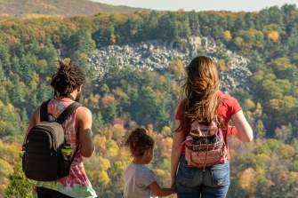 A white woman, a Black man, and a young girl face away from the camera and look at the view of trees changing color for Fall and several hills