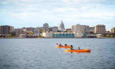 A family kayaks on Lake Monona in front of Monona Terrace. The photo is zoomed out to show both kayaks with two people on each and the Madison skyline in the background.