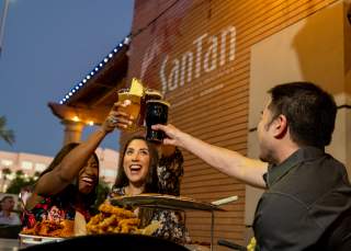 Two women and one man cheers beers on SanTan Brewing patio with food.