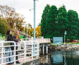 Two people pause along Nanaimo’s waterfront walkway, leaning on a railing and enjoying the view of the calm water and nearby park.