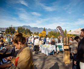 People browsing stalls at Queenstown craft market with autumnal trees and the Remarkables mountain range in the background