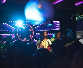 Person singing and playing guitar on a stage with colourful lights and neon sign