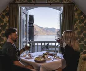 A cosy dinner setting with a couple enjoying wine and a delicious meal, overlooking stunning lake and mountain views through open balcony doors.