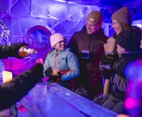 A family of four enjoying drinks in an ice bar, surrounded by illuminated ice sculptures and glowing purple and blue lighting. They are dressed warmly in coats, hats, and gloves, smiling and sharing a joyful moment.