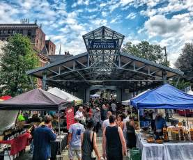 A photo of the Lexington Farmers Market at the Fifth Third Pavilion. People can be seen shopping at different vendor’s tents. The sky above the pavilion is a beautiful blue color with clouds peppered throughout.