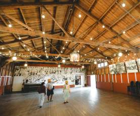 Three people explore a spacious wooden hall with exposed beams, string lights, and historic photos on the walls.