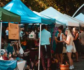 Visitors browse vendor stalls under colourful tents at a busy outdoor market set among tall trees.