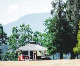 A small red pavilion sits among tall trees with mountains in the background on a sunny day.
