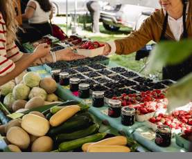 A vendor hands a basket of strawberries to a customer at a farmers’ market, with fresh produce and jars of preserves on display.