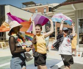 Three people walking on the street, each holding a different flag over their heads. From left to right- Pride flag, lesbian flag, and trans flag.