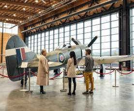 Visitors admire a vintage aircraft displayed in a well-lit museum with large windows and red ropes marking the exhibit area.