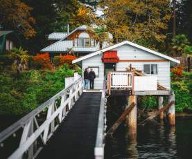Two people stand on a narrow walkway leading to a small oceanfront house on stilts above the water, surrounded by trees and fall foliage.