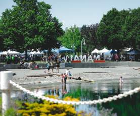 People gather along a waterfront park in Nanaimo (Maffeo Sutton Park), with children wading in the water and market tents and the ‘Nanaimo’ sign in the background.