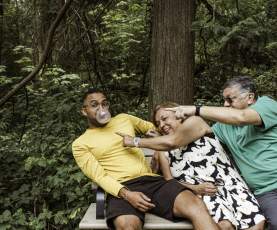 A man in a yellow shirt blows a bubblegum bubble while sitting on a forest park bench as an older couple beside him laugh and reach across playfully to try to pop it.