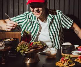 A person in a striped shirt uses chopsticks to pick up food from a table filled with tacos, greens, and drinks.