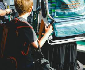 A child wearing a backpack boards a ferry while holding onto an adult carrying a folded beach chair.