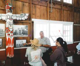A guide explains historical photos to two visitors next to a carved totem pole inside a rustic wooden hall.