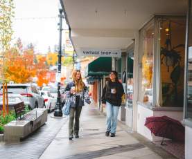 Two people stroll past shops on a rainy autumn day in downtown Nanaimo, holding coffee as colourful fall trees line the street.