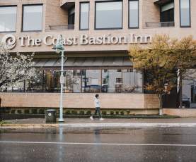 A person walks past The Coast Bastion Inn on a rainy day, with wet pavement and trees lining the street.