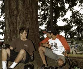 Two men sitting leaning on a tree with a bike.