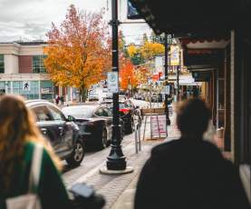 People walk along a downtown Nanaimo street with shops, parked cars, and vibrant autumn trees lining the road.