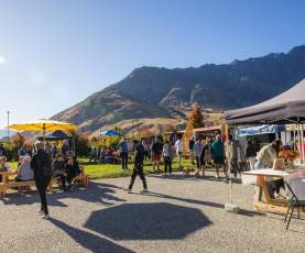 People browsing stalls and relaxing under colourful umbrellas at the Remarkables Market, with mountains in the background on a sunny day.