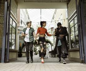 Three friends walk side by side, laughing in a bright, covered shopping walkway lined with storefronts.