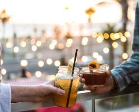 Pacific City in Huntington Beach, California, featuring two people holding handcrafted cocktails on an outdoor patio, with warm string lights and a coastal sunset atmosphere in the background.