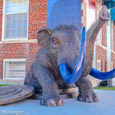 A sculpture of a mastodon, featuring prominently in in front of the Muskegon Museum of History and Science with buildings in the background. The sculpture is marked by realistic textures and a striking contrast between its gray body and blue tusks.