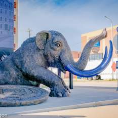 A large sculpture of a mastodon with blue tusks is placed outside the Lakeshore Museum Center. The creature is crouched on a circular base, and a "Visit Muskegon" logo is seen in the corner. The background includes a brick building and a delivery truck.