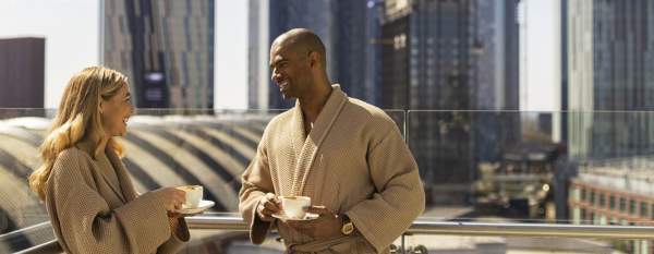 Couple on hotel balcony in dressign gowns drinking coffee with views over Manchester