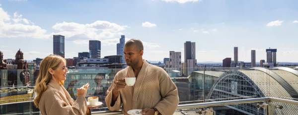 Couple in dressing gowns drinking coffee on Hotel Balcony overlooking Manchester