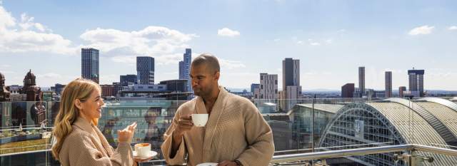Couple in dressing gowns drinking coffee on Hotel Balcony overlooking Manchester