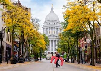 A man carries a giant "M" across a street with the Capitol building in the background