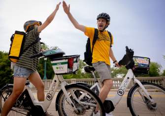 A white man and an Asian woman high five while riding electric bikes on a clear day in Madison