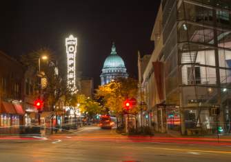 A view of State Street looking toward the Capitol building at night. Street lights are lit up and the light from a passing by car can be seen in the foreground.