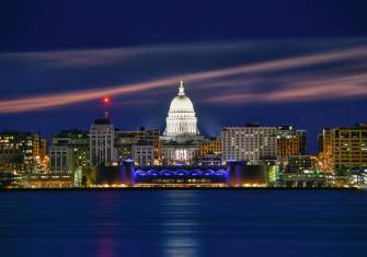 Night time view of downtown Madison from Lake Monona. The Capitol is lit up with bright white lights, and the Monona Terrace has blue lights along the arches. One of the buildings has lights on in a heart shape.