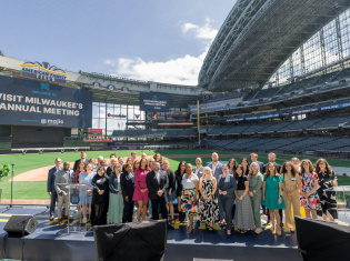 Group photo of attendees at Visit Milwaukee's Annual Meeting, posed on a stage at American Family Field (Milwaukee Brewers stadium) with stadium seating and banners visible in the background.