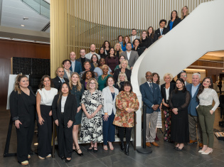 Large group of professionals posing on a curved staircase inside a modern building with wood slat walls and glass railings.