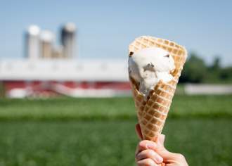 A hand holding up a cone of ice cream in front of a barn
