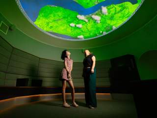 2 women stand in the centre of a dark circular room looking up at a bright projection of the Humber Estuary in vivid blue and green.
