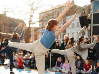 Three dancers stand on one leg in Trinity Square with a crowd surrounding them