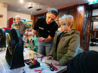 A chikd in a green coat with their face painted is stood next to a woman in a black t shirt who is working on a model wind turbine fan on the table in front on her.
