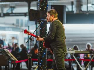 A singer stands on stage with a guitar in Trinity Market, Hull. People sit at tables in the background.