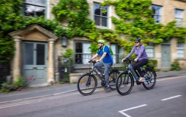 Two cyclists riding bikes through picturesque Charlbury, passing stone cottages covered with greenery