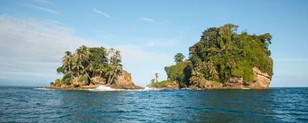 This image shows two small, lush, rocky islands covered in tropical vegetation, surrounded by the ocean. The islands are close to each other, separated by a narrow water channel. Palm trees and dense greenery dominate the landscape, and the sky is mostly clear with a few clouds, indicating a bright, sunny day.