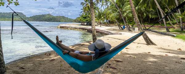 A person in a wide-brimmed hat relaxes in a turquoise hammock strung between two palm trees on a white sand beach. In the background, calm, clear turquoise water meets the shore, with a rocky area visible to the right. Beyond the water, lush green hills rise under a partly cloudy sky. More palm trees line the beach on Mamey Island