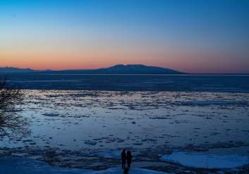 People stand at the edge of Cook Inlet with Mount Susitna across the water at dusk.