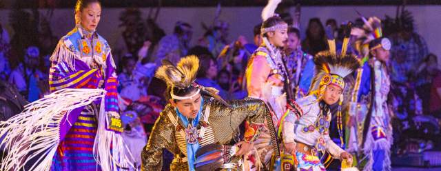 Indigenous performers in traditional outfits perform on a large stage, swathed in blue and orange light
